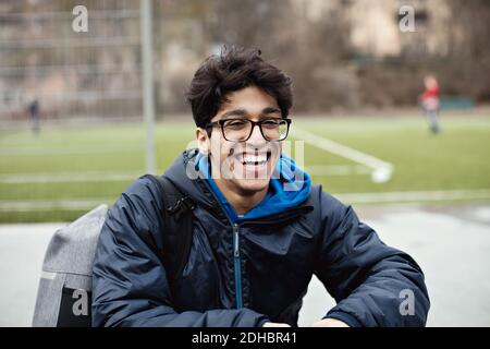 Fröhlicher junger Mann in warmer Kleidung und Brille beim Sitzen Gegen Fußballplatz in der Stadt Stockfoto