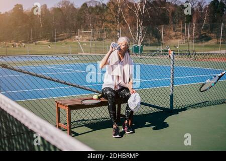 Müde ältere Frau, die Wasser trinkt, während sie auf der Bank sitzt Tennisplatz Stockfoto