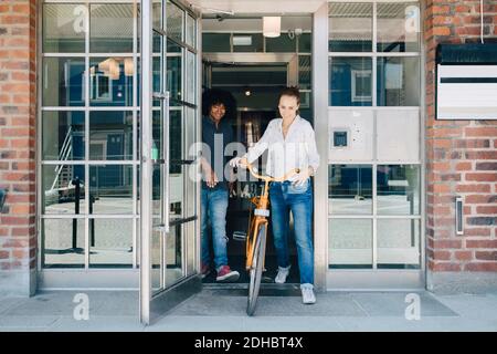Die ganze Länge der lächelnden Geschäftsfrau hält Fahrrad beim Gehen mit Kollege durch die Tür im Büro Stockfoto