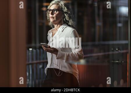 Durchdachte Geschäftsfrau mit Kopfhörern, während sie das Mobiltelefon bei benutzt Büro Stockfoto