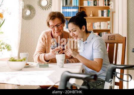 Lächelnde junge Sozialarbeiterin, die ältere Rentnerinnen unterstützt Mit Smartphone am Tisch im Pflegeheim Stockfoto
