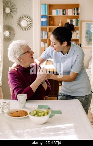 Lächelnde Rentnerin hält die Hände einer jungen Freiwilligen Während des Gesprächs im Pflegeheim Stockfoto