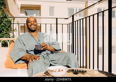 Glücklicher reifer Mann, der Kaffee hat, während er auf dem Sofa sitzt Balkon Stockfoto