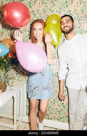 Porträt einer lächelnden jungen Frau, die im Stehen mit Ballons spielt Vom Mann gegen Tapete zu Hause beim Abendessen Teil Stockfoto