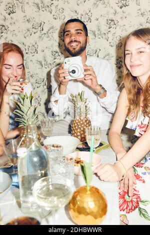 Lächelnder junger Mann, der die Kamera hält, während er inmitten weiblicher Freunde sitzt Zu Hause während der Dinner-Party Stockfoto