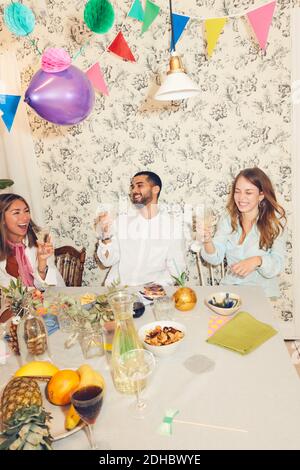 Fröhliche junge Freunde feiern beim Sitzen am Esstisch während Dinnerparty Stockfoto