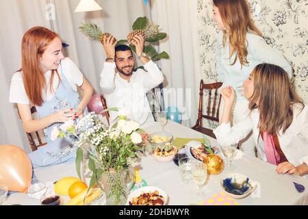 Spielerischer Mann, der Ananas hält, während er inmitten weiblicher Freunde sitzt Zuhause während der Dinner-Party Stockfoto