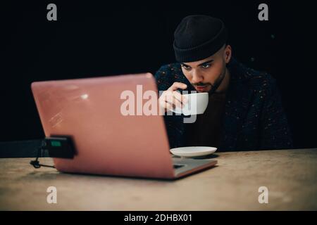 Selbstbewusst junge männliche Hacker trinken Kaffee, während Blick auf Laptop Am Schreibtisch im Büro Stockfoto