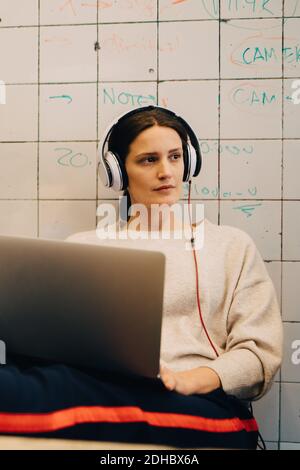 Junge Computerhackerin, die mit Kopfhörern zuhört, während sie mit sitzt Laptop gegen Fliesenwand im Kreativbüro Stockfoto