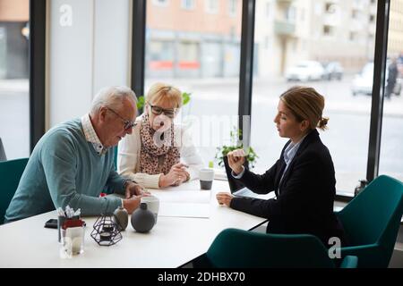 Älterer Mann mit Frau, die das Eigentumsdokument unterschreibt, während sie mit sitzt Makler im Immobilienbüro Stockfoto