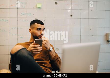 Junger männlicher Programmierer schaut weg, während er mit Smartphone sitzt Gegen die Wand im Büro Stockfoto