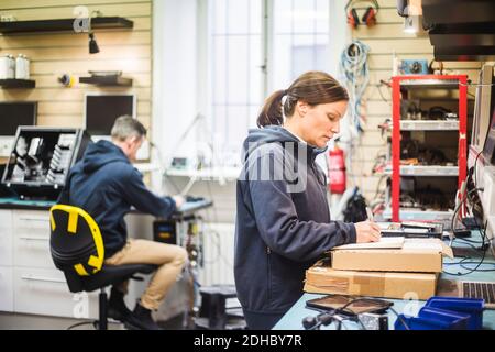 Seitenansicht des Technikers beim Schreiben auf das Buch, während der Kollege arbeitet Im Hintergrund beim Workshop Stockfoto