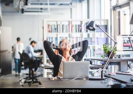 Reife Geschäftsfrau sitzt mit den Händen hinter dem Kopf am Schreibtisch in Büro Stockfoto
