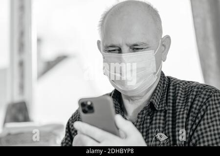 Schwarz-Weiß-Bild eines älteren Mannes mit Schutzhandschuhen Und chirurgische Maske während der Bedienung eines Pro-Smartphone in der Öffentlichkeit Platz Stockfoto