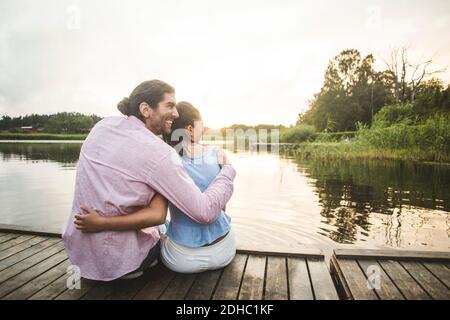 Rückansicht eines lächelnden Paares, das mit den Armen um sich herum sitzt Steg über dem See bei Sonnenuntergang Stockfoto