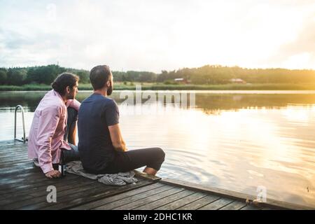 Männliche Freunde, die beim Sitzen auf dem Steg den See betrachten Sonnenuntergang Stockfoto