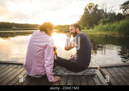 Lächelnde männliche Freunde reden, während sie auf dem Steg über dem See sitzen Bei Sonnenuntergang Stockfoto