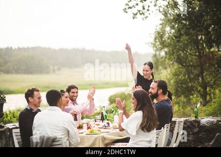 Frau mit mittlerem Erwachsenen steht mit erhobenem Arm, während Freunde applaudieren Am Tisch während der Dinner-Party im Hinterhof Stockfoto