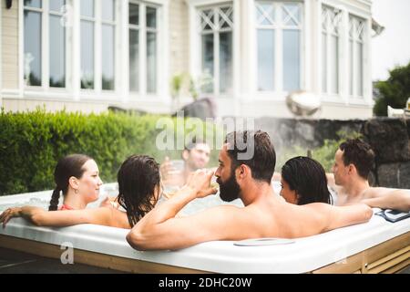 Mittlerer Erwachsener Mann, der Alkohol trinkt, während er mit Freunden in genießt Wannen-Whirlpool am Wochenende Stockfoto