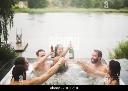 Unbeschwerte männliche und weibliche Freunde genießen Getränke im Whirlpool Gegen den See während des Wochenend-Kurzurlaubs Stockfoto