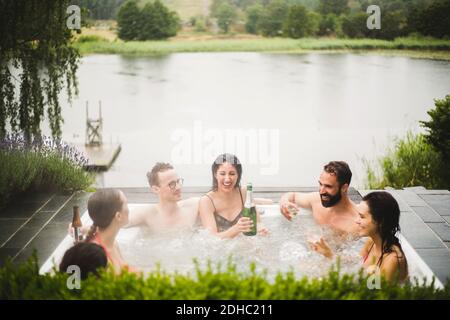 Fröhliche männliche und weibliche Freunde genießen Getränke im Whirlpool Gegen den See während des Wochenend-Kurzurlaubs Stockfoto