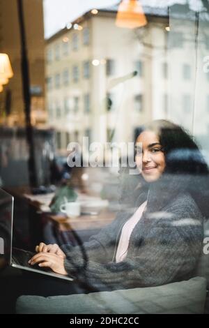 Portrait von lächelnden jungen weiblichen professionellen sitzen mit Laptop gesehen Durch das Fenster im Büro Stockfoto