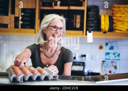 Ältere Verkäuferin lächelt, während sie ein braunes Ei aus der Pappe pflückt Feinkostgeschäft Stockfoto