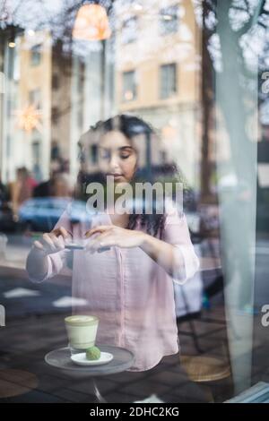 Junge Influencerin fotografiert Tee und Snack am Tisch Smartphone durch Fenster gesehen Stockfoto