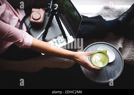 High-Angle-Ansicht der jungen weiblichen Influencer trinken Matcha-Tee Beim Erstellen von Vlog im Kreativbüro Stockfoto