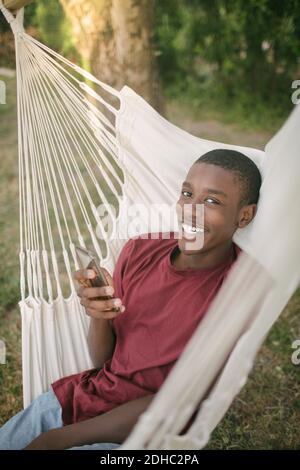 Porträt eines lächelnden Jungen mit Mobiltelefon, während er sich zurücklehnend Hängematte im Hinterhof Stockfoto