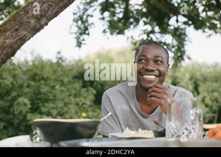 Lächelnder Mann mit mittlerem Erwachsenenalter, der wegschaute, um am Tisch zu Mittag zu essen Während Gartenparty Stockfoto