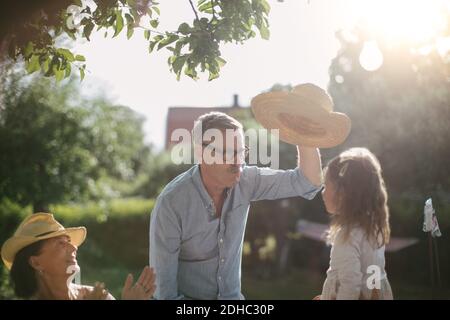 Frau klatscht und schaut auf einen älteren Mann, der Strohhut hält Über Enkelin im Hinterhof während sonnigen Tag Stockfoto