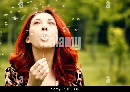 Schönes Mädchen in einem modischen Leoparden Print Jacke zwischen den grünen Bäumen. Sommer romantische Stimmung. Spaziergang im Park. Eine junge rothaarige Frau bläst einen Löwenzahn weg. Stockfoto