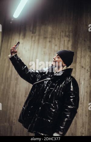 Junge Frau trägt schwarze glänzende Jacke während der Aufnahme Selfie mit Handy gegen Wand in U-Bahn Stockfoto