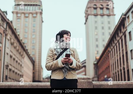 Fröhliche junge Frau, die wegschaut, während sie das Handy dagegen hält Gebäude in der Stadt im Winter Stockfoto