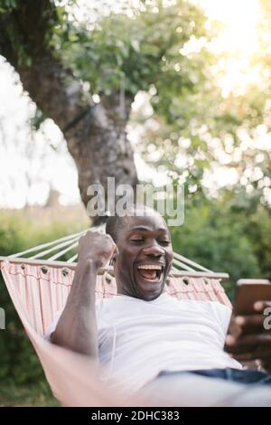 Ein fröhlicher Mann mittleren Erwachsenen, der beim Entspannen auf das Handy schaut Auf Hängematte im Hinterhof Stockfoto