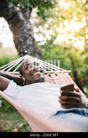 Lächelnder Mann mit mittlerem Erwachsenenalter, der Musik über das Mobiltelefon hört Entspannen in der Hängematte im Hinterhof Stockfoto