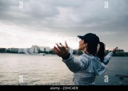 Weibliche Sportlerin, die mit den vom Meer ausgestreckten Armen gegen den Himmel steht In der Stadt Stockfoto