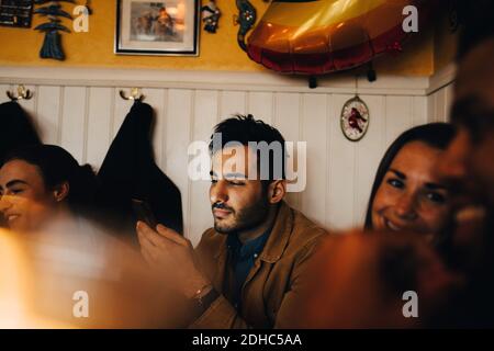 Junger Mann, der Handy benutzt, während er unter Freunden sitzt Restaurant Stockfoto