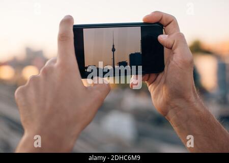 Zugeschnittenes Bild eines jungen Mannes, der den Fernsehturm über ein Smartphone fotografiert Bei Sonnenuntergang Stockfoto