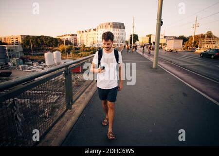 Ganze Länge des jungen Mannes mit Handy beim Gehen Auf dem Fußweg bei der Brücke in der Stadt Stockfoto