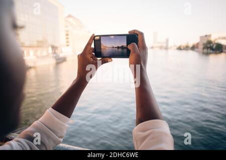 Zugeschnittenes Bild einer jungen Frau, die den Fluss über das Mobiltelefon fotografiert In der Stadt Stockfoto