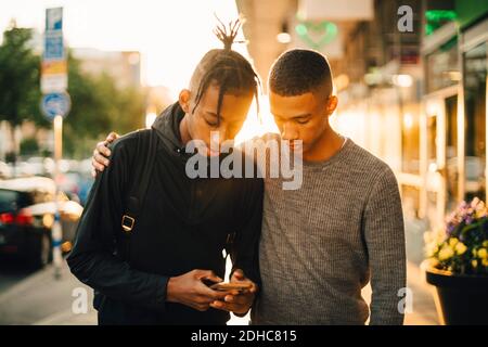 Teenager-Junge mit Handy, während Sie von einem Freund auf Straße in der Stadt bei Sonnenuntergang Stockfoto