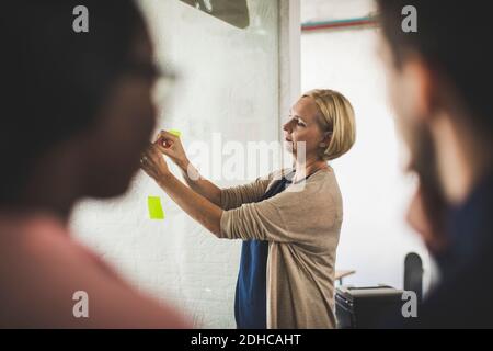 Unternehmer, die eine Kollegin anschaut, die eine Klebenote auf Glas klebt Im kreativen Büro Stockfoto