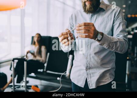 Mittelteil eines Geschäftsmannes, der im Stehen verwickelte Ohrhörer hält abflugbereich des flughafens Stockfoto