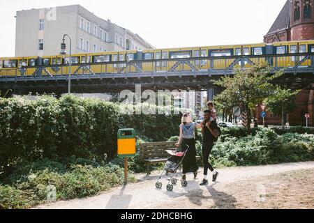 Junger Mann, der Tochter auf den Schultern trägt, während Frau Baby schiebt Kinderwagen am Fußweg gegen die Eisenbahnbrücke Stockfoto