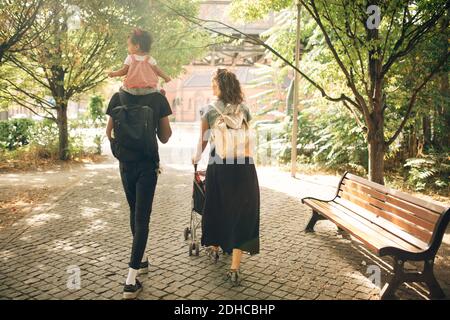 Rückansicht des Mannes, der Tochter auf den Schultern trägt, während Frau Kinderwagen auf Fußpfad schieben Stockfoto