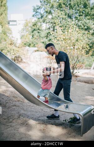Junger Mann hält die Hände der Tochter, die auf der Rutsche läuft Spielplatz Stockfoto