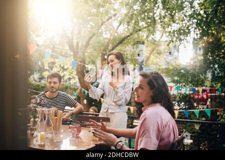 Lächelnde junge Frau Videokonferenzen über Smartphone, während Freunde Sitzen auf dem Balkon während der Party Stockfoto