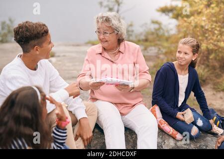 Glückliche Großmutter, die dem Enkel ein Geschenk gibt, während sie mit Enkeltöchtern sitzt Im Park während des Picknicks Stockfoto
